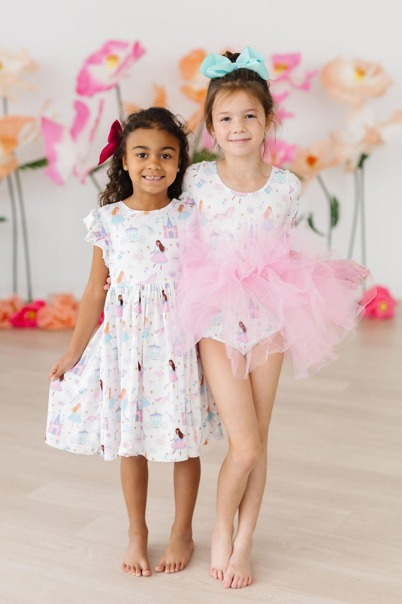 Two happy girls showcasing the Fairy Princess 3/4 Sleeve Tutu Leotard and a matching dress by Mila & Rose, posing together with playful smiles, surrounded by colorful flowers. A delightful duo ready for dance adventures!