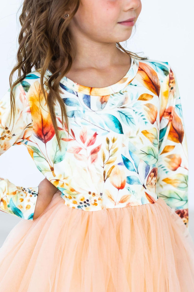 Girl wearing a fall watercolors long sleeve tutu dress featuring colorful leaf print top and peach tulle skirt.