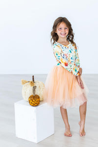 Girl wearing fall watercolors long sleeve tutu dress with peach skirt and autumn leaf print top near woven pumpkin decor.