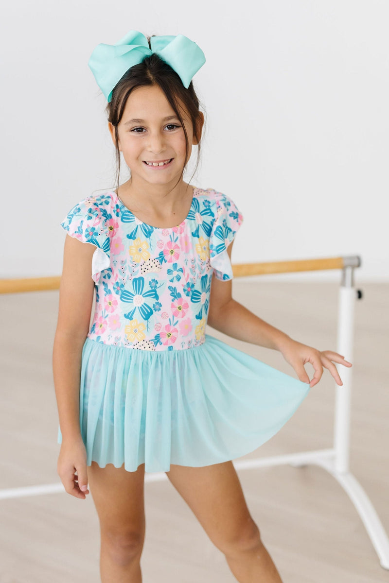 A smiling girl wearing a Floral Garden Short Sleeve Twirl Leotard for Girls with a matching bow, ready for dance class.