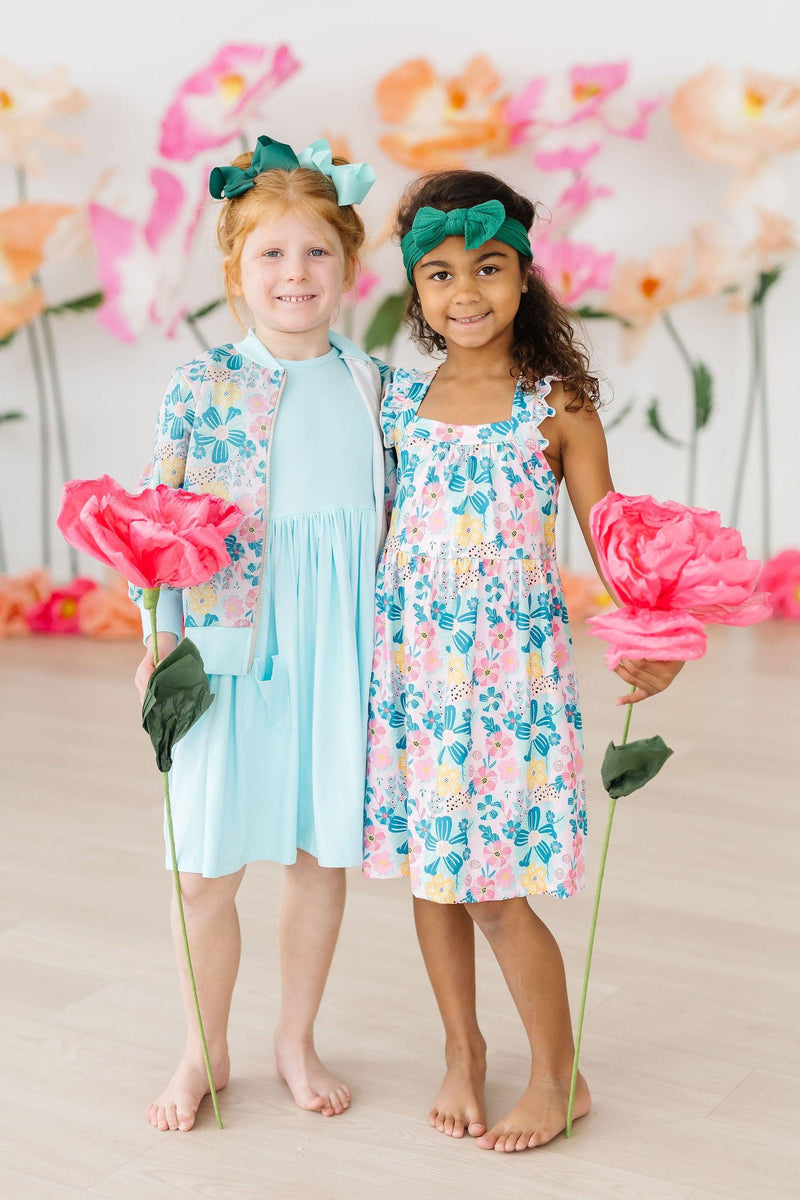Adorable duo wearing the Floral Garden Shimmer Jacket and matching dresses from Mila & Rose, perfect for twirling in style amid a backdrop of oversized flowers.