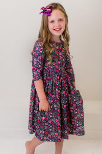 Flower Farm 3/4 Sleeve Pocket Twirl Dress worn by a smiling girl in navy floral print with a purple bow.