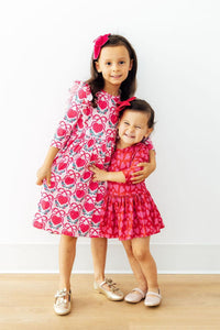 Two girls hugging in pink heart-print 3/4 sleeve ruffle twirl dresses with matching bows. Perfect for school or play.