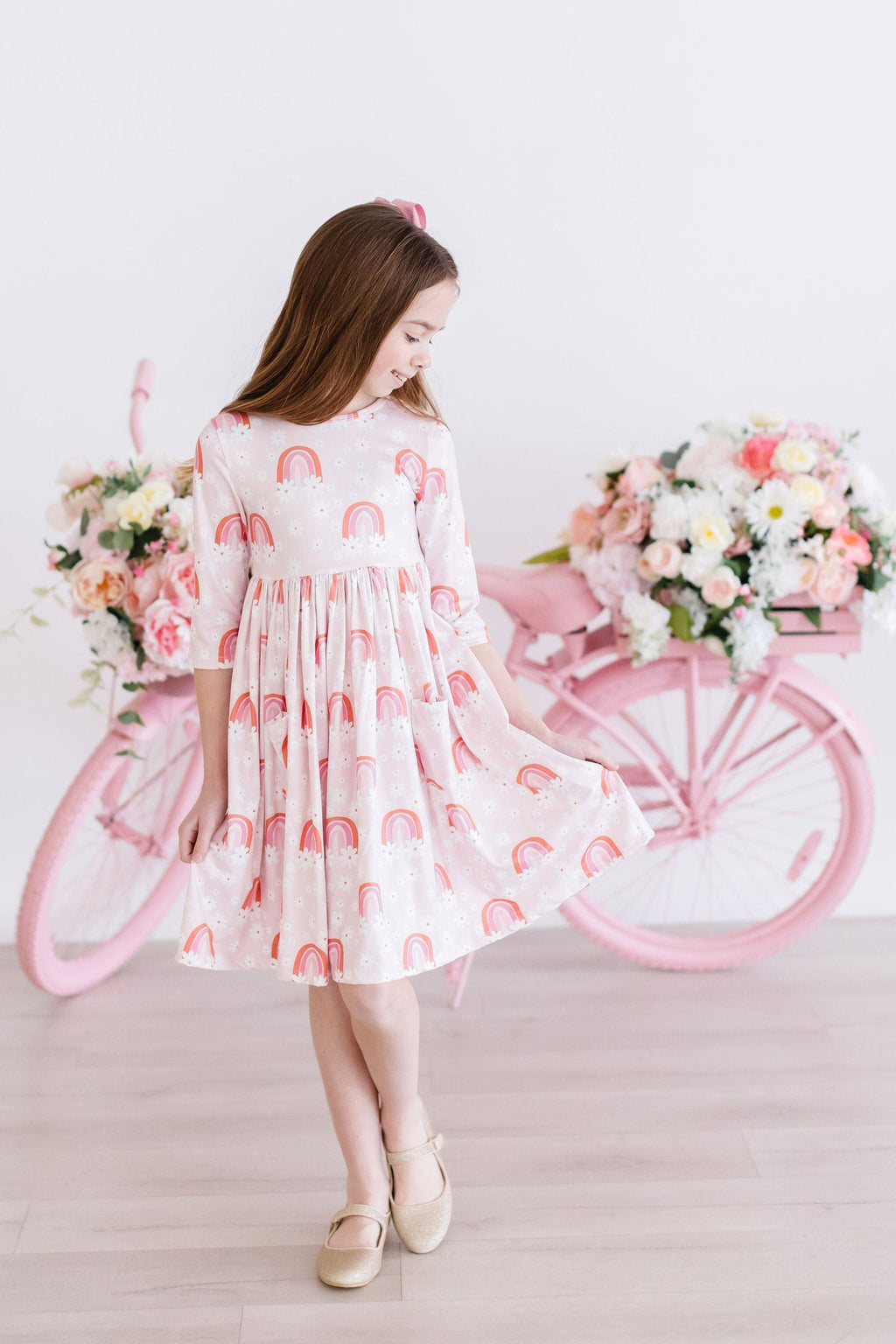 A young girl twirling happily in the Fun & Flowers 3/4 Sleeve Pocket Twirl Dress, featuring colorful rainbows, set against a charming backdrop with pink bicycles and floral decorations.