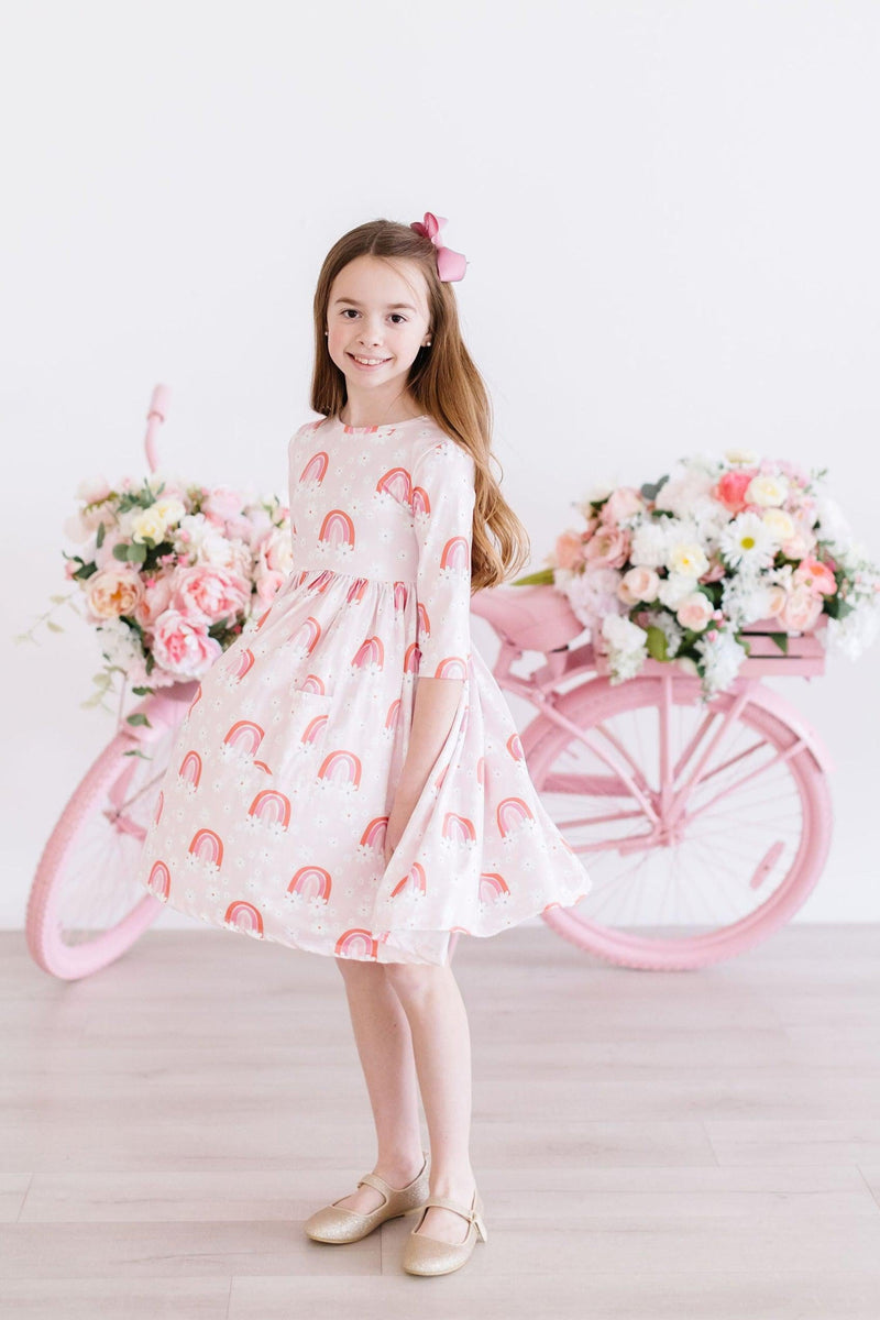 Charming young girl posing in the Fun & Flowers 3/4 Sleeve Pocket Twirl Dress adorned with playful rainbows, complemented by a pink bicycle and beautiful floral arrangements in the background.