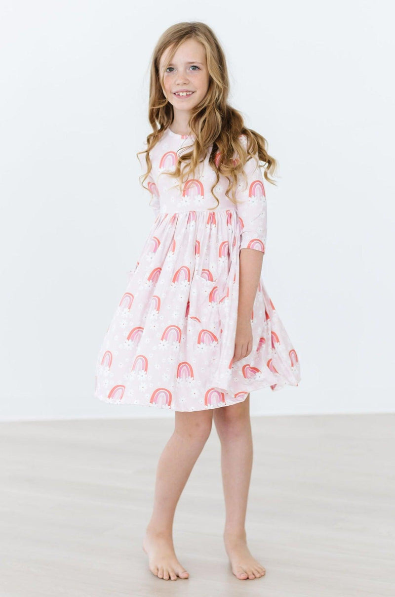 A joyful child wearing the Fun & Flowers 3/4 Sleeve Pocket Twirl Dress by Mila & Rose, showcasing a playful rainbow print. Perfect for twirling and adventures, this dress combines style and comfort for every young girl's wardrobe.