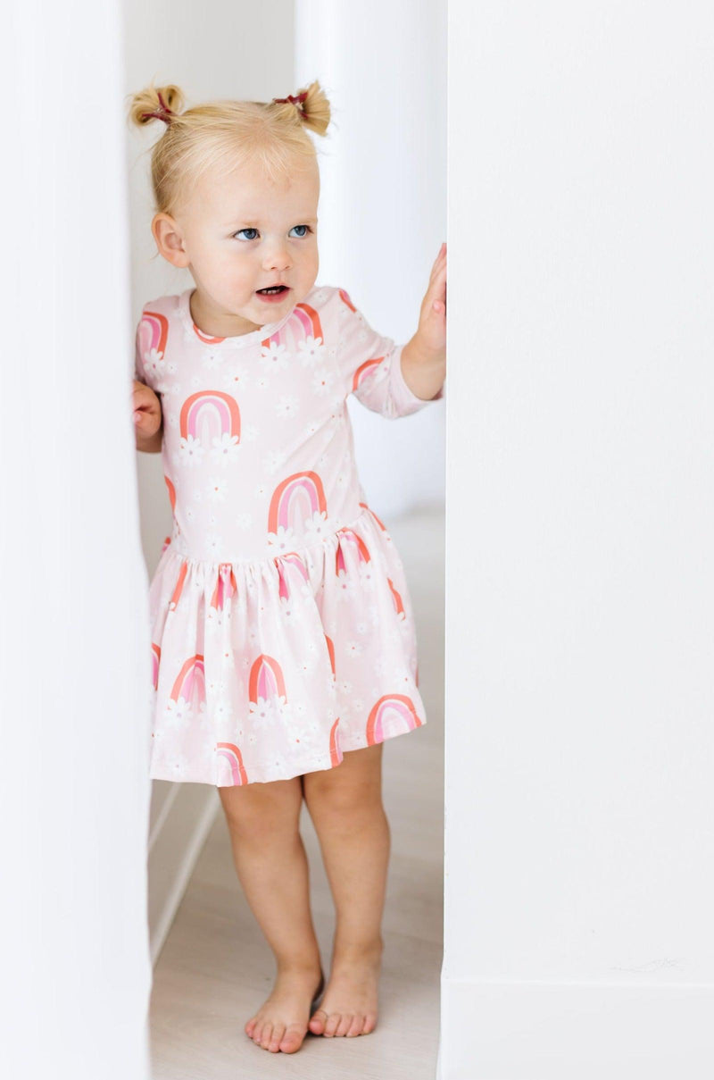 Cute toddler peeking playfully while dressed in the Fun & Flowers Twirl Bodysuit by Mila & Rose®, showcasing a delightful pink and rainbow pattern perfect for twirling adventures.