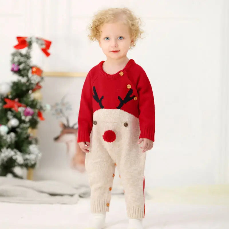 A toddler wearing the Rudy Reindeer Knit Children's Winter Romper in red, smiling in a decorated setting.