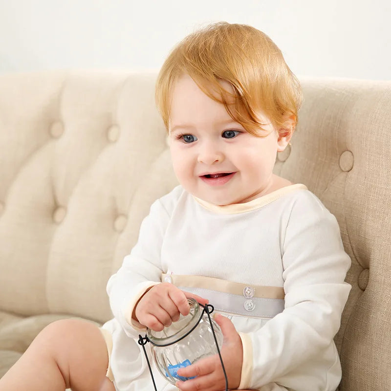 Smiling baby girl in a white long-sleeve formal dress with beige trim sitting on a tufted sofa holding a glass jar.