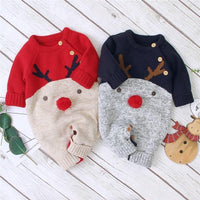 Two baby knit one-piece outfits laid flat on a white wood plank background. One is red with a beige lower section and dark brown reindeer antlers; the other is navy with a gray lower section and brown antlers.