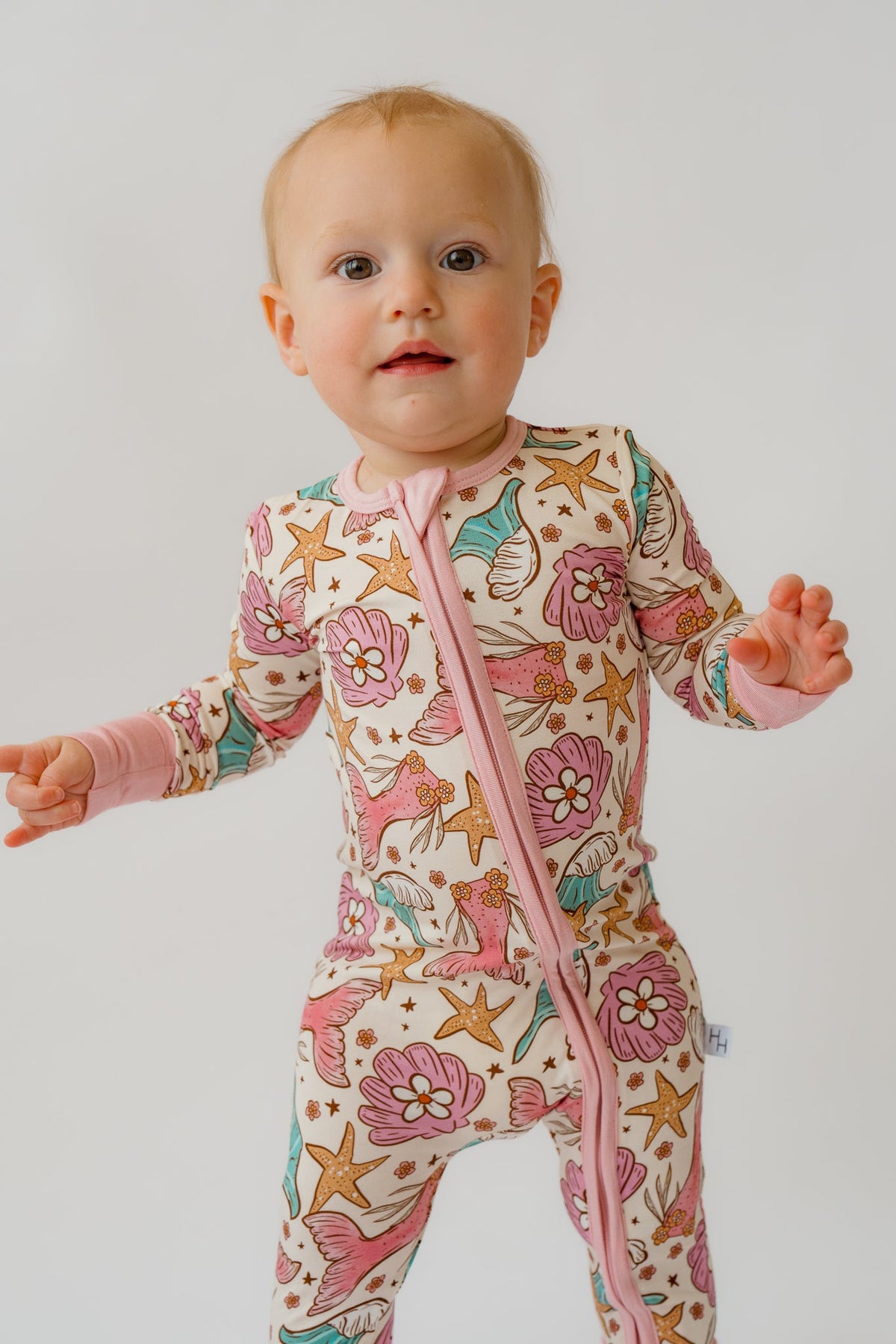Baby wearing cream mermaid print onesie with pink tails, teal fins, and diagonal zipper with pink guard