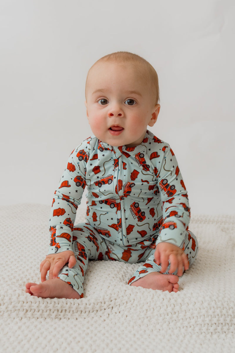 Baby sitting on white blanket wearing light blue fire engine patterned bamboo sleeper with zipper