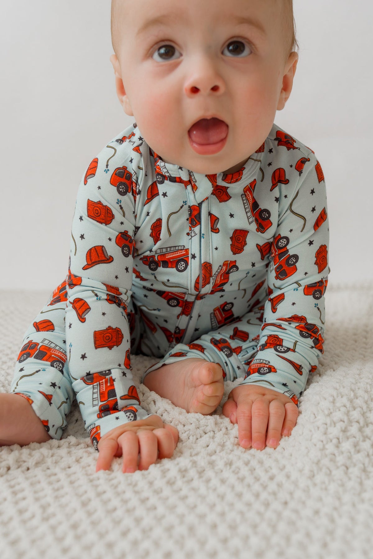 Baby crawling on white rug in light blue fire truck print onesie with red trucks and black stars