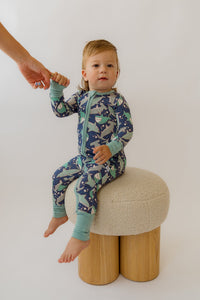 Toddler sitting on a stool wearing a blue shark-print convertible bamboo onesie with teal trim.