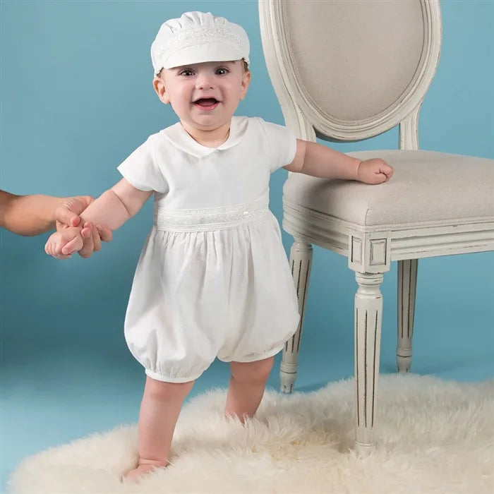Smiling baby boy in a white special occasion romper with turn-down collar and matching cap, standing by a chair on a white rug.