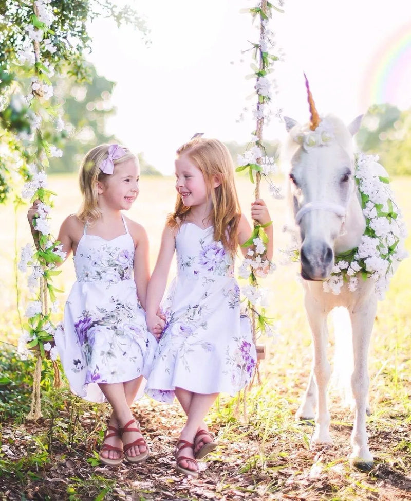 Two little girls in matching 'Oh Suzannah' Floral Party Dresses, smiling happily while swinging near a unicorn.