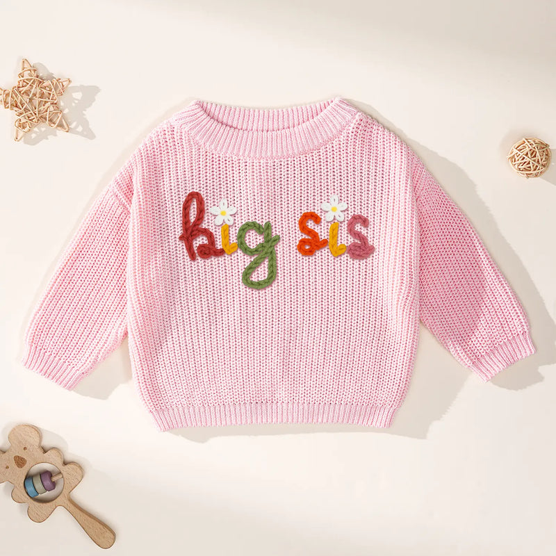 Light pink chunky knit crewneck sweater with colorful 'big sis' embroidery and white flowers on cream background