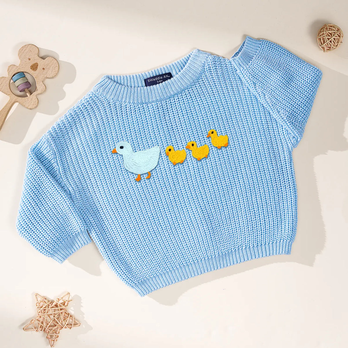 Baby blue pullover with ribbed cuffs showing mother duck embroidery near wooden bear rattle