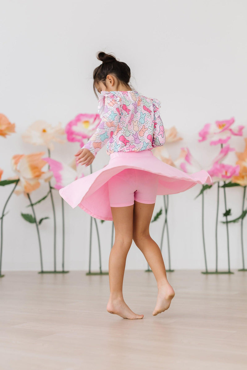 A young girl spins joyfully in a pink skirt, showcasing the Hangin with My Peeps L/S Ruffle Tee by Mila & Rose, featuring playful bunny prints. Background adorned with colorful flowers, perfect for showcasing cheerful children's fashion.