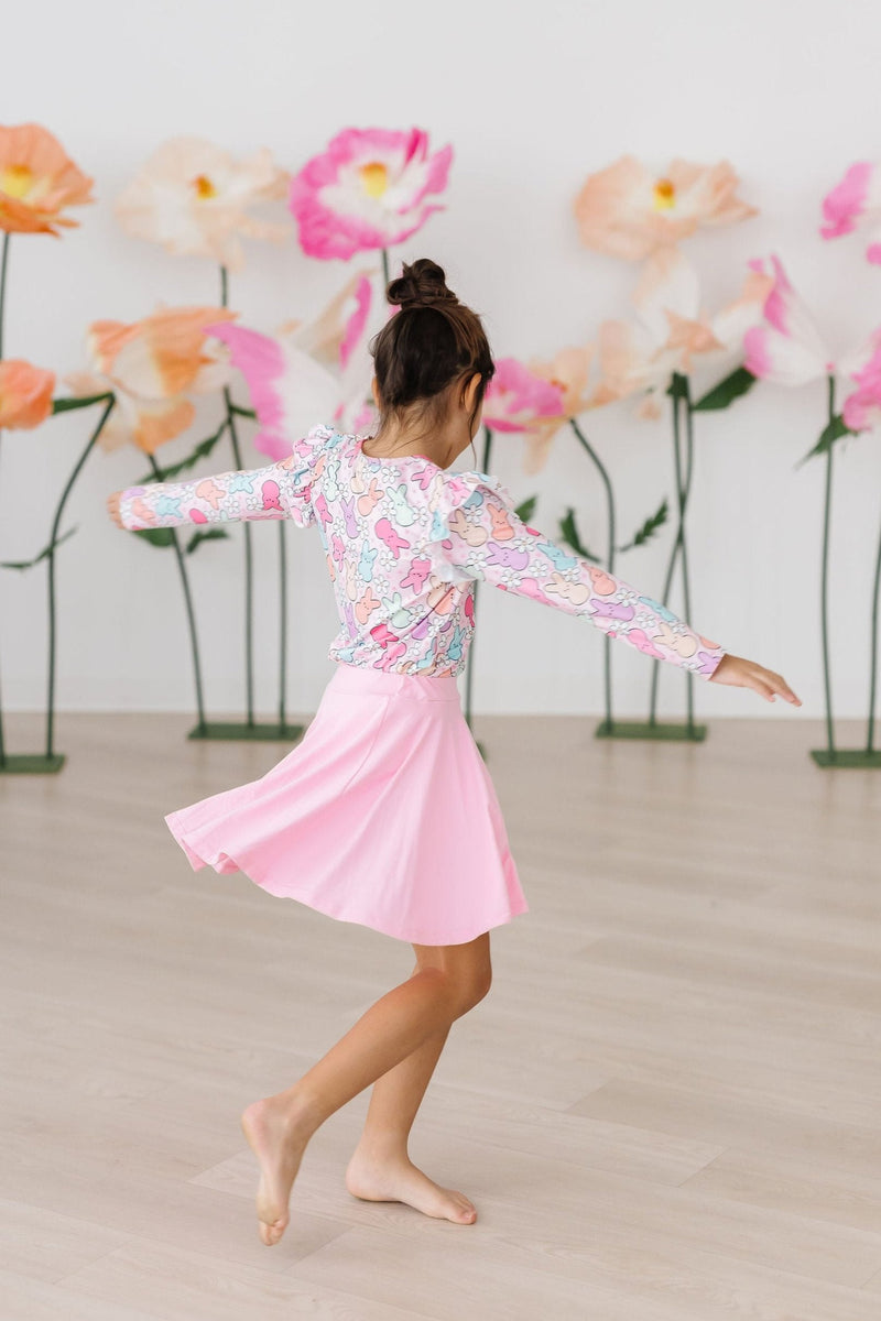 A girl twirls in a pink skirt while wearing the Hangin with My Peeps L/S Ruffle Tee by Mila & Rose, characterized by vibrant bunny and flower patterns. The image captures the essence of fun and playful style for kids.