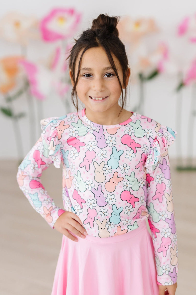 A smiling girl poses confidently in the Hangin with My Peeps L/S Ruffle Tee by Mila & Rose, highlighted by a fun design of bunnies and flowers. She wears a pink skirt, embodying the spirit of playful fashion for young girls.