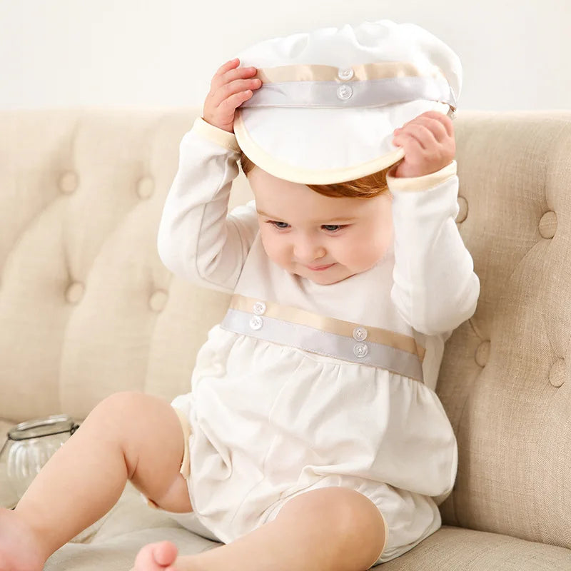 Toddler boy in white formal romper with beige trim and matching cap, sitting on tufted sofa for special occasions.