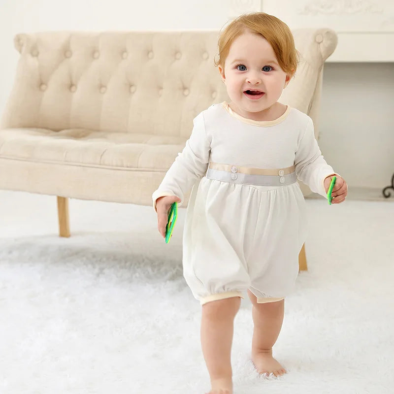 Smiling toddler boy wears a white formal romper with a decorative button waistband, perfect for special occasions.