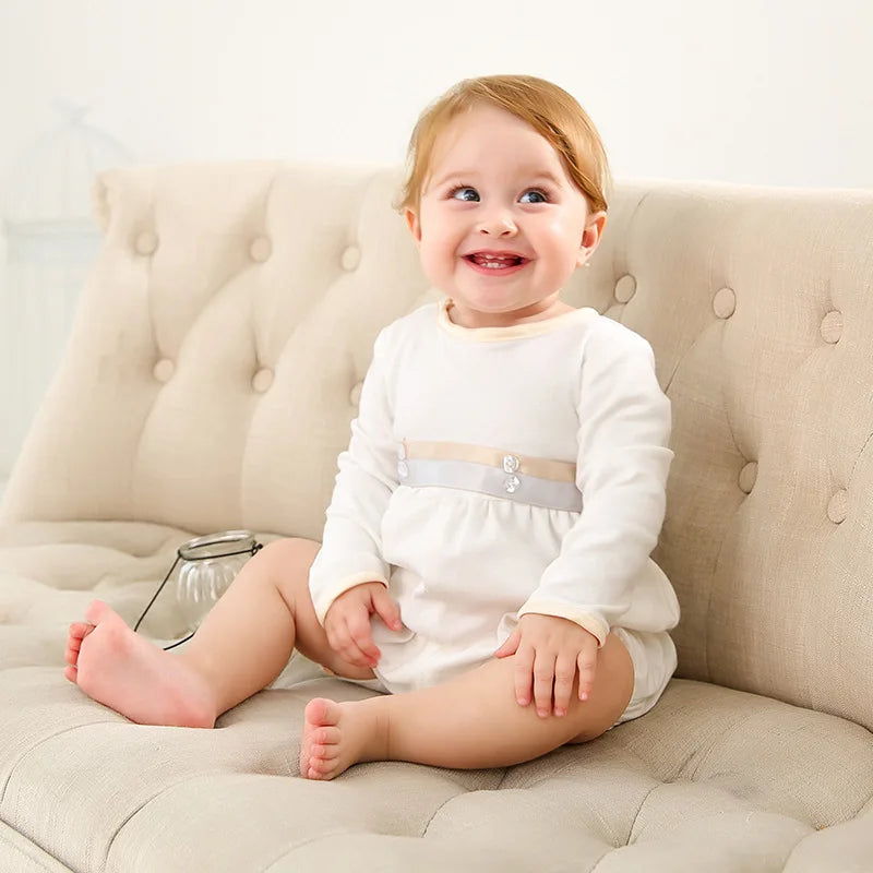 Smiling baby girl wearing a white formal romper with decorative ribbon and buttons, sitting on a tufted beige sofa.
