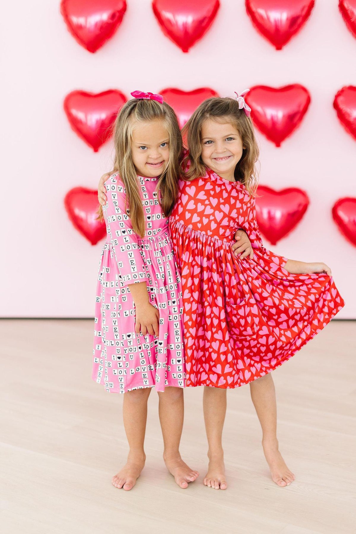 Two girls pose with heart balloons wearing a pink I Love You print and a red heart print 3/4 sleeve pocket twirl dress.
