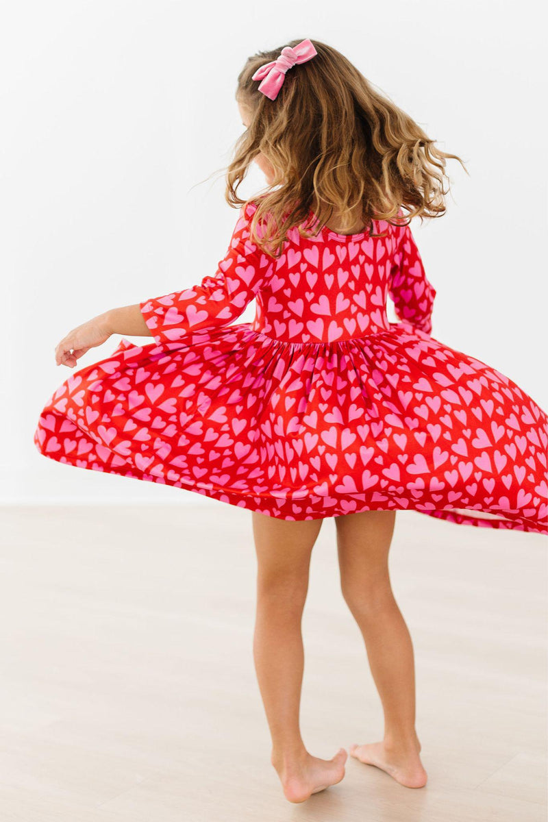 Girl twirling in a red and pink heart print 3/4 sleeve dress. Features a full skirt, scoop back, and soft fabric for play.