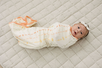 Baby swaddled in GOTS certified organic cotton blanket with boho floral print and peach trim, lying on quilted beige mat.