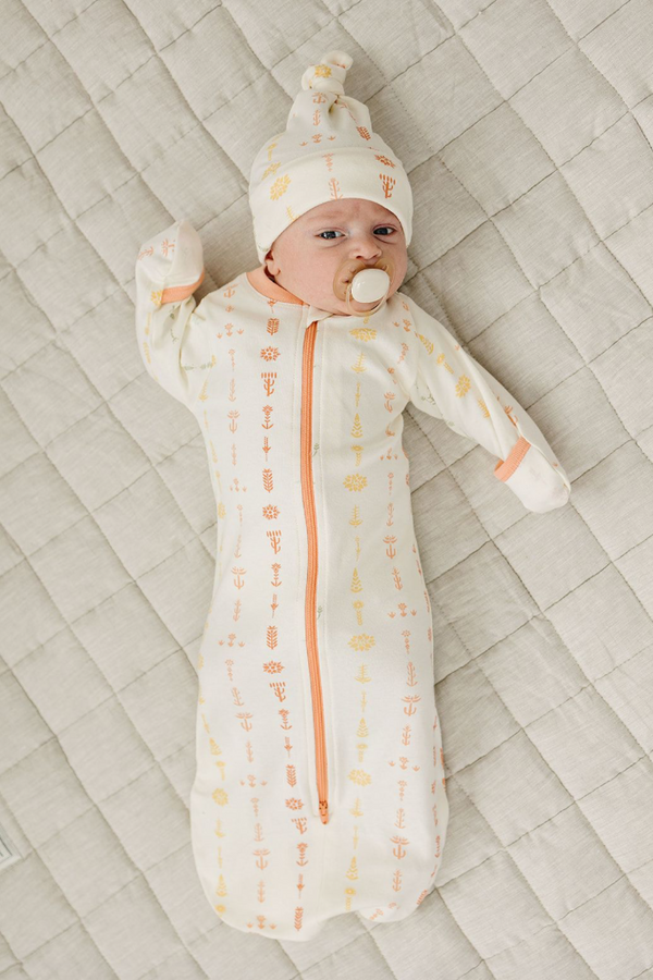 Baby wearing GOTS certified organic cotton sleepsack with boho floral print and matching hat, lying on quilted bedding.