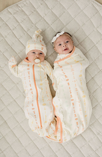 Two babies on a quilted mat; one wears a gOTS certified organic cotton sleepsack with boho floral print and orange zipper, the other is swaddled in matching fabric.