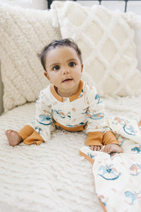 Baby wearing GOTS certified organic cotton playsuit with rocking horse print and orange trim, sitting on textured cream blanket.