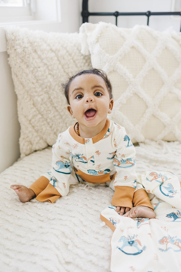 Happy baby wearing GOTS certified organic cotton playsuit with rocking horse print and amber trim, sitting on textured white bedding.