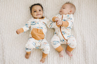 Two babies lying on a knit blanket wearing GOTS certified organic cotton playsuits with a rocking horse print and rust-colored pocket.