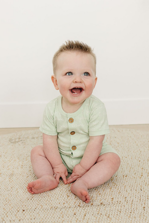 Happy baby sitting on a rug wearing a desert green organic cotton short sleeve romper with "I Love You" text and wooden buttons.