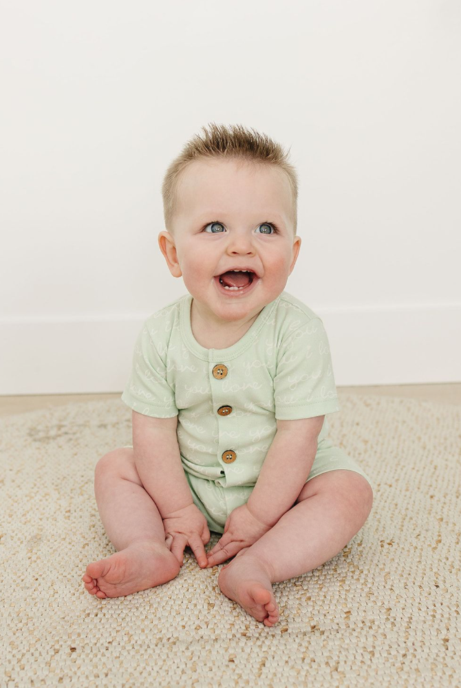 Happy baby sitting on a rug wearing a desert green organic cotton short sleeve romper with "I Love You" text and wooden buttons.