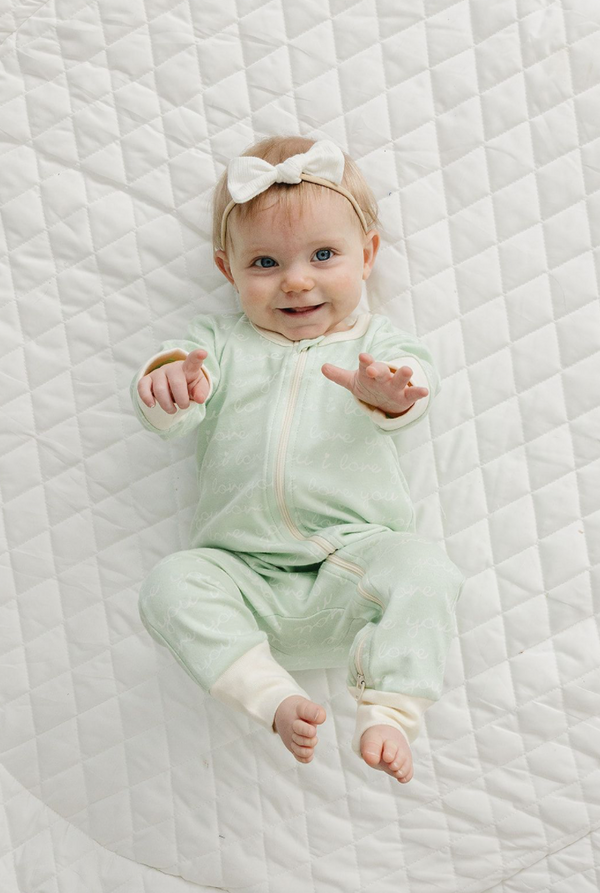 Smiling baby wears GOTS certified organic cotton zippy romper in desert green with "I love you" print on white quilt.