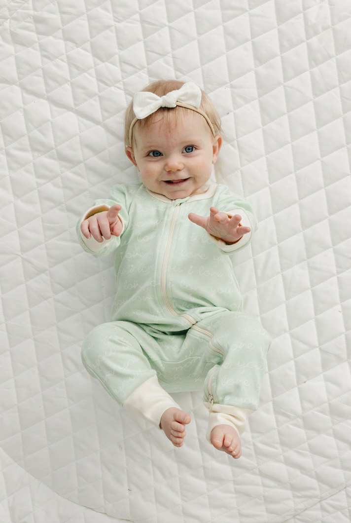 Smiling baby wears GOTS certified organic cotton zippy romper in desert green with "I love you" print on white quilt.