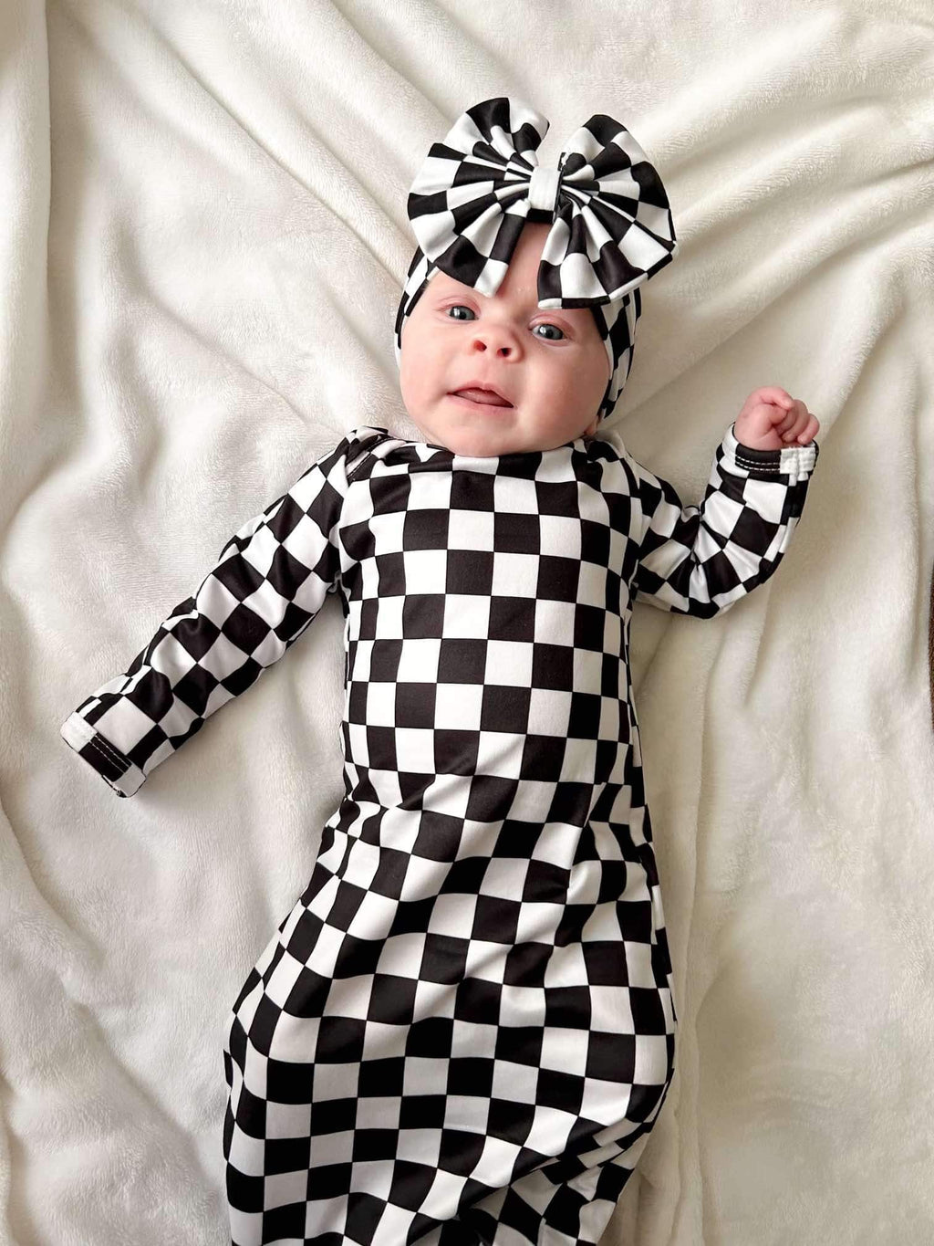 Adorable baby lying down in a Checkered Knotted Baby Gown Set with Headband. This stylish knotted baby gown set features a classic black and white checkered design, showcasing its comfort and chic appeal.