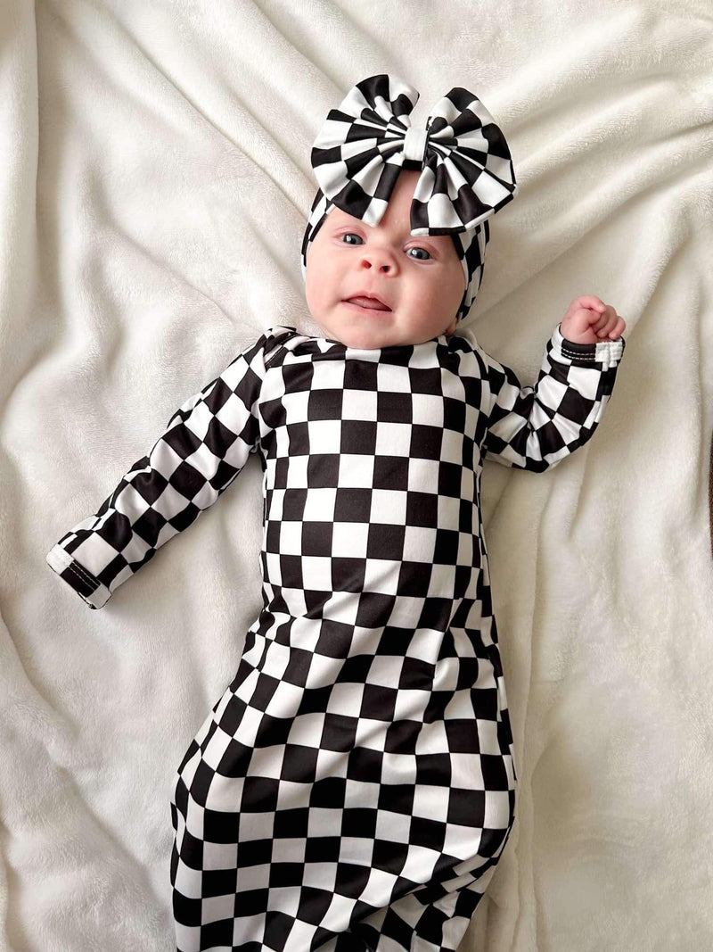 Adorable baby lying down in a Checkered Knotted Baby Gown Set with Headband. This stylish knotted baby gown set features a classic black and white checkered design, showcasing its comfort and chic appeal.