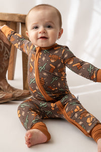 Baby wearing a brown Highland cow and cactus print bamboo loverall with orange zipper trim.