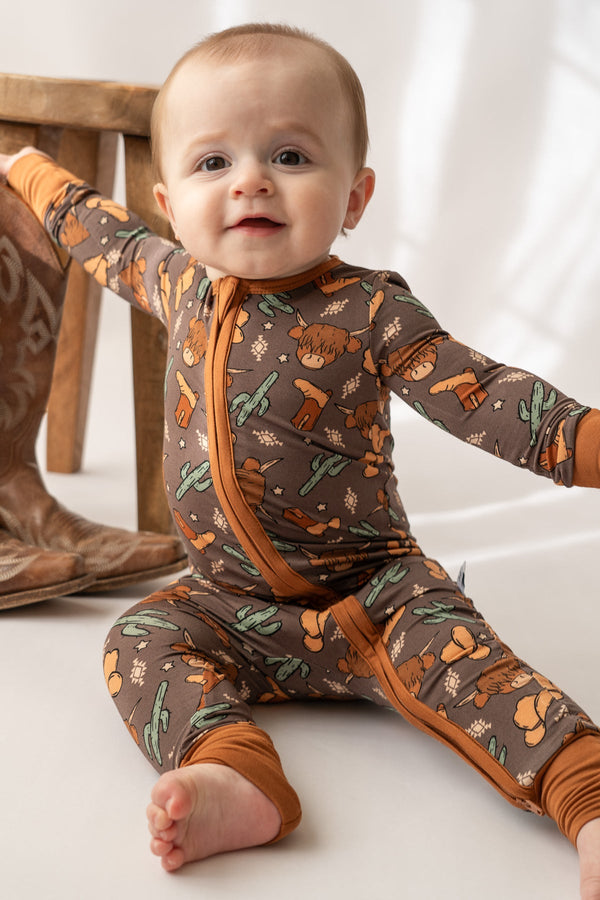 Baby wearing a brown Highland cow and cactus print bamboo loverall with orange zipper trim.
