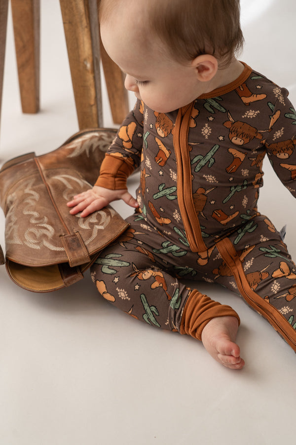 Baby wearing a brown Highland cow and cactus print bamboo loverall, touching a cowboy boot.