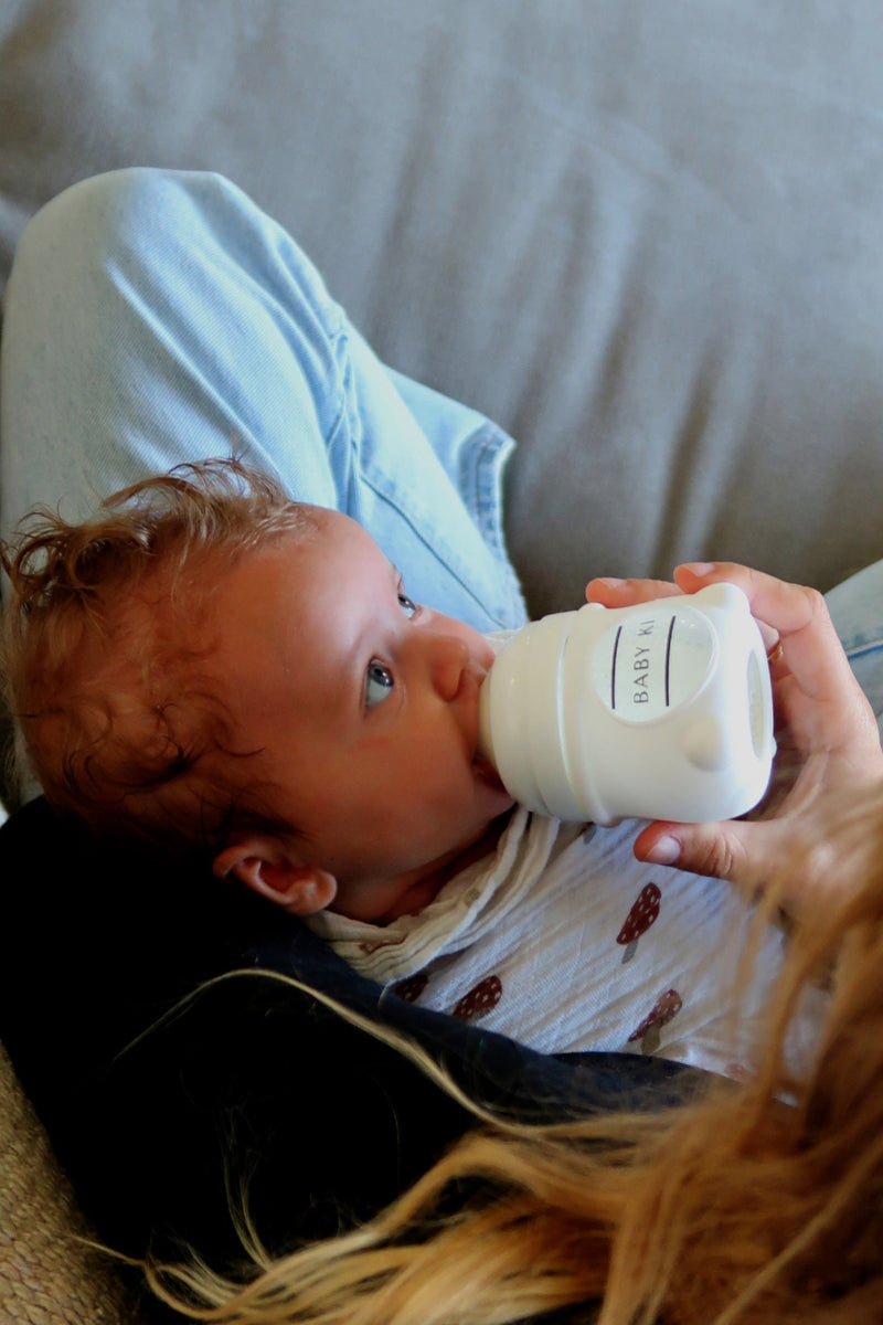 Baby drinking from a bottle held by an adult on a couch