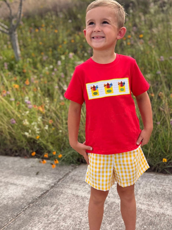 Little boy in red shirt with crayon embroidery and yellow gingham shorts outdoors.