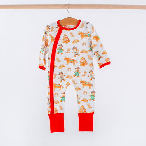 White organic cotton baby zip-up pajama with red trim, featuring a festive cookie cowboy print with horses, trees, and boots.