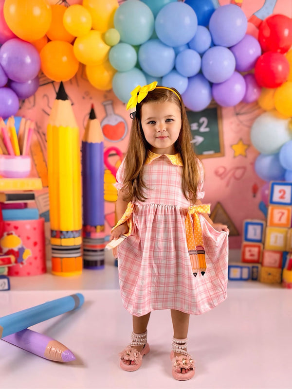 The Girls Back To School Pencil Twirl Dress on a happy girl posing in front of vibrant balloons and school decor, showcasing the cute plaid design and bow details.