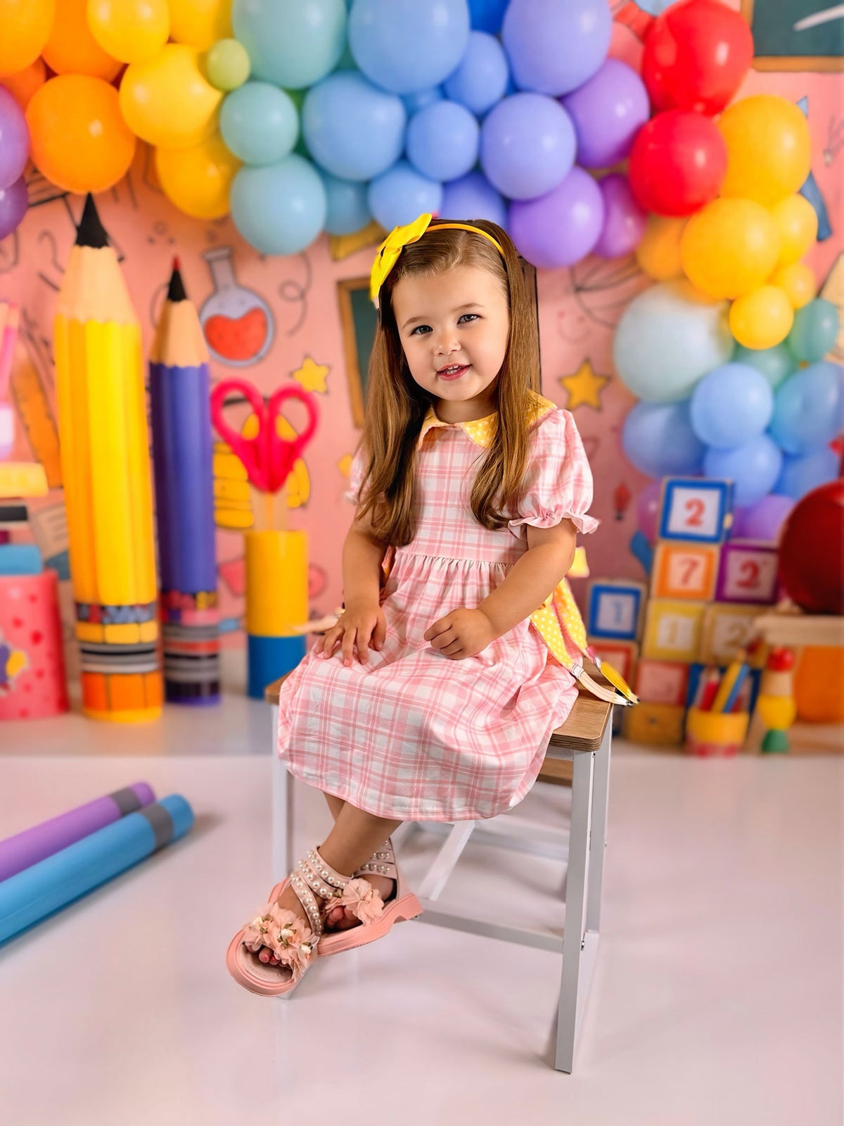 Girl sitting on a chair wearing the Girls Back To School Pencil Twirl Dress, showcasing the dress's charming bow appliques against a colorful backdrop.